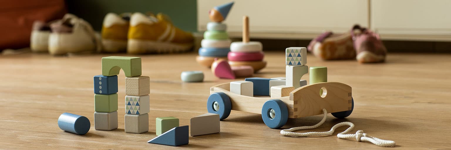 A wooden pull-along cart, stacking rings, and building blocks arranged on a wood floor by an entryway