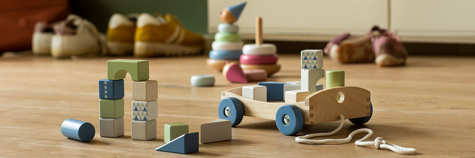 A wooden pull-along cart, stacking rings, and building blocks arranged on a wood floor by an entryway